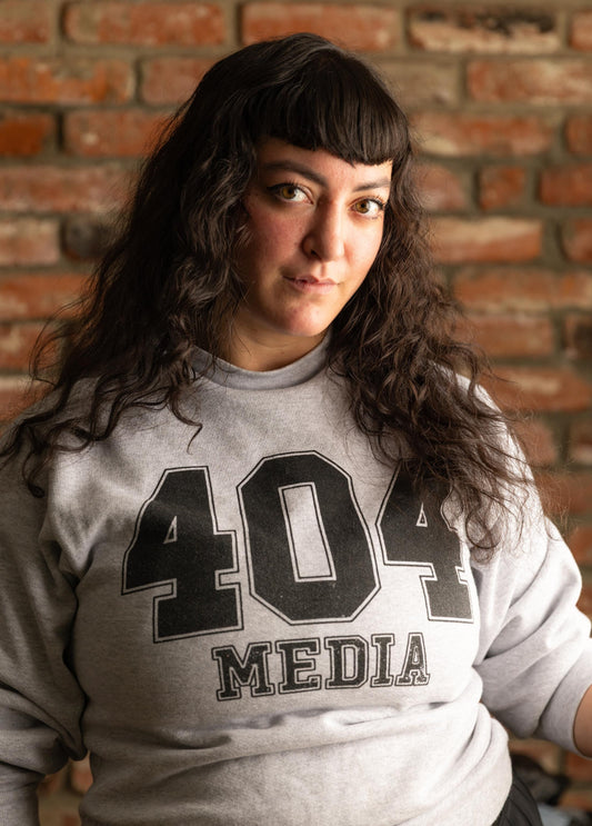 Person wearing a gray sweatshirt with '404 Media' text against a brick wall.